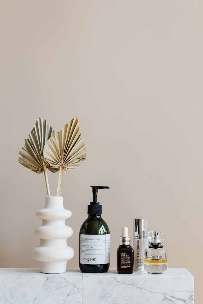 Assorted bottles with cosmetics products placed with creative geometric vase with artificial leaves on white marble shelf against beige wall in modern bathroom interior