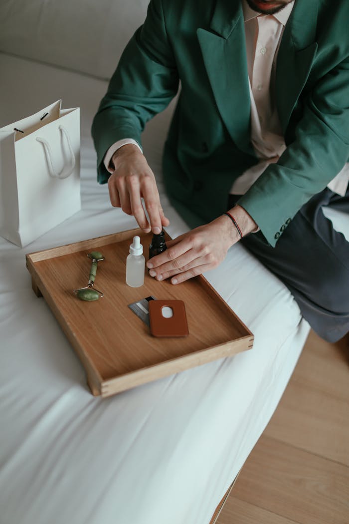 High angle of crop anonymous male in fashionable outfit sitting on comfortable bed and testing various cosmetic products after online shopping